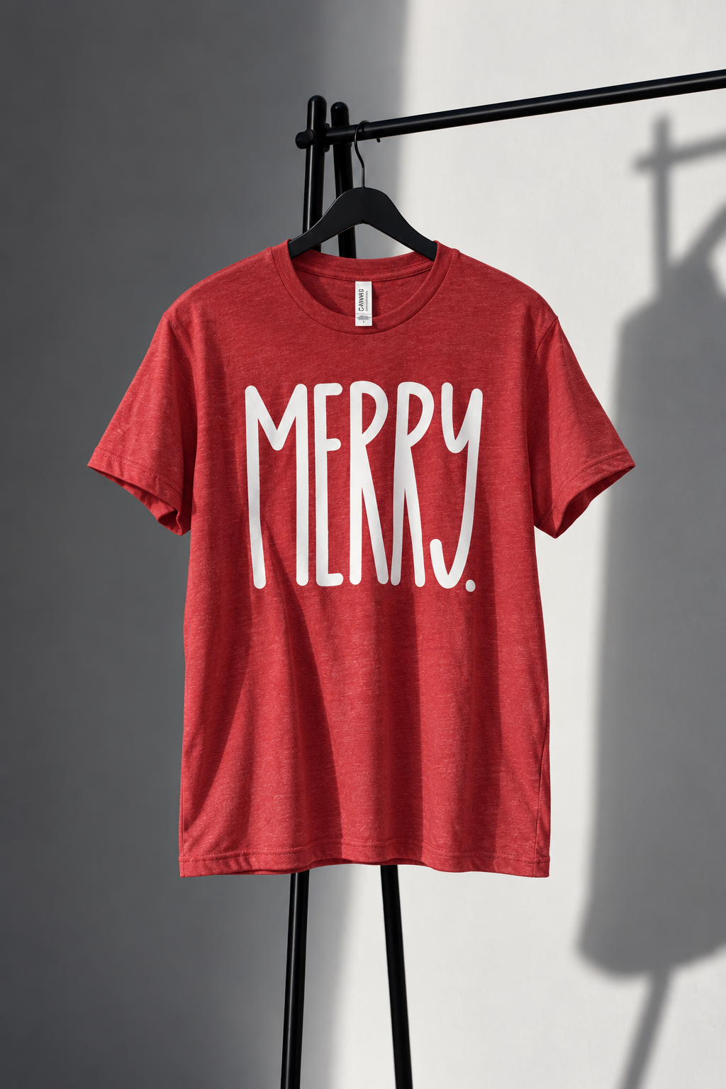 Red Bella Canvas tee with tall white MERRY lettering on black studio rack