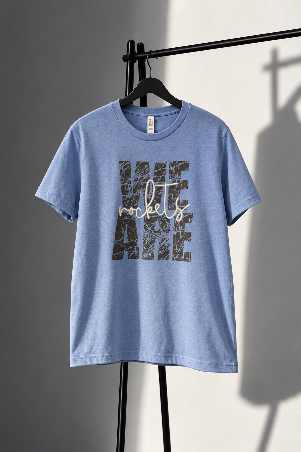Light blue Bella+Canvas medium tee with distressed WE ARE lettering and cream rockets script